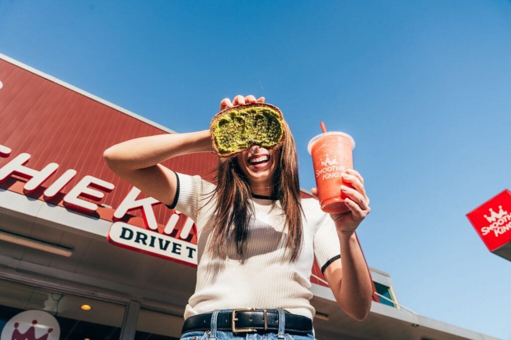 A woman holding an Avocado Loaded Toast and a Smoothie.